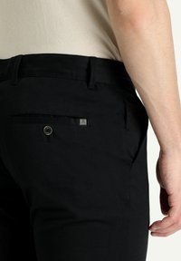 Pantalones negros de material tejido con corte recto. Las características incluyen un bolsillo trasero con un botón y una pequeña etiqueta de logo.