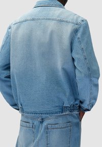 Vista posteriore di una persona che indossa una giacca di jeans azzurra chiara e jeans coordinati con tasche e orlo elastico sulla giacca.