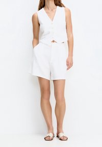 Un crop top en lin blanc avec un décolleté en V profond, comportant six boutons. Associé à un short blanc taille haute, les deux ayant des poches avant et une coupe décontractée.