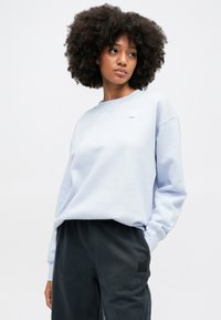 Világoskék pulóver kerek nyakkivágással és hosszú ujjakkal, kis folttal díszítve. Sötétszürke melegítőnadrággal párosítva.