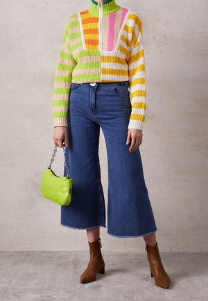 Personne portant un pull tricoté rayé multicolore, un jean bleu large et court, des bottines marron, tenant un petit sac à main vert citron avec une bandoulière en chaîne.
