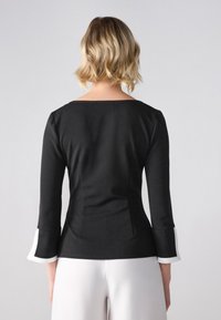 Haut ajusté noir avec manches trois-quarts, doté de poignets blancs. Tissu lisse avec une légère brillance et un col arrondi.