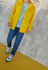 Person iført en gul regnjakke, lysblå skjorte, blå jeans og hvid-grønne sneakers, som går på et gult gulv med en tekstureret grå væg i baggrunden.