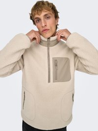 Pull polaire beige avec col montant et fermeture éclair à moitié. Comprend une poche frontale avec un rabat marron clair et deux poches latérales zippées. Texture douce.