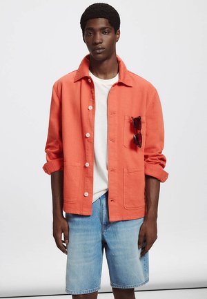 Jeune homme portant une veste corail orange à boutons par-dessus un t-shirt blanc et un short en jean bleu clair, avec des lunettes de soleil dans la poche de la veste.
