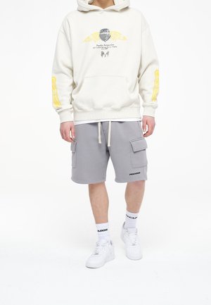 Sweat à capuche crème avec motif graphique et accents jaunes, associé à un short cargo gris avec poches. Baskets blanches et chaussettes montantes avec logo.