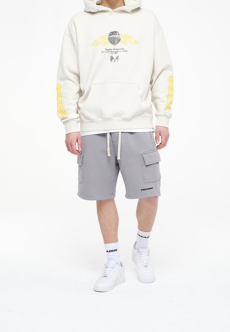 Sweat à capuche crème avec motif graphique et accents jaunes, associé à un short cargo gris avec poches. Baskets blanches et chaussettes montantes avec logo.