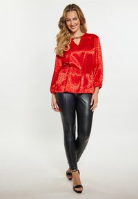 Rote satinierte Bluse mit Tiermotiv, mit einem Schlüsselloch-Ausschnitt und langen Ärmeln, kombiniert mit schwarzen Lederleggings und schwarzen Absatzschuhen.