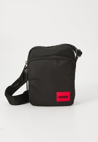 Black nylon crossbody bag featuring a red rectangular logo patch, zippered compartments, adjustable strap, and a textured surface.