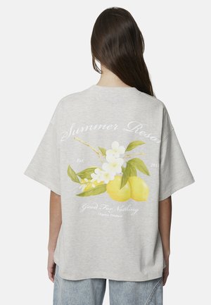 Osoba z długimi brązowymi włosami, ubrana w szarą, oversize'ową koszulkę z grafiką cytryny i białego kwiatu oraz ozdobnym tekstem na plecach.