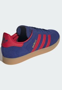 Blå ruskindsneakers med røde striber og hæltab, gummisål og "GAZELLE" skrevet i guld på siden.