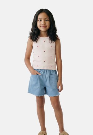 Jeune fille aux cheveux bouclés foncés portant un haut blanc sans manches avec des cœurs rouges, un short bleu avec des poches et des chaussures beiges à enfiler, debout