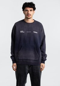 Tmavě námořnický oversized svetr vyrobený z bavlněné směsi, má přední kapsu, kulatý výstřih a tři tištěné loga v bílé barvě.