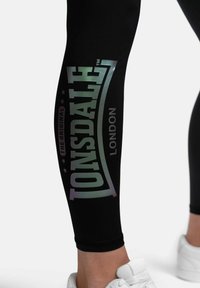 Black leggings featuring a glossy, iridescent logo stating "Lonsdale London" along the side. Smooth texture with a fitted design.