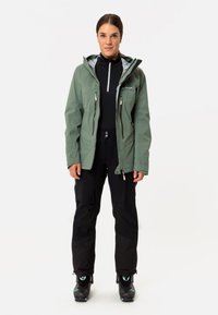Grüne wasserfeste Jacke mit Kapuze, vorderen Reißverschlüssen und grauem Innenfutter, kombiniert mit schwarzen Hosen und Stiefeln mit türkisfarbenen Akzenten.