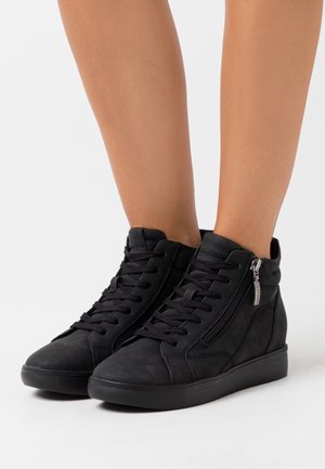 Baskets montantes noires avec lacets et fermeture éclair latérale portées sur des jambes nues sur un fond blanc uni.