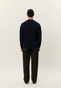 Άνδρας που φοράει μπλε σκούρο πουλόβερ, μαύρο τζιν και μαύρο καπέλο του μπέιζμπολ, στέκεται αντικρίζοντας ένα απλό ανοιχτό φόντο.