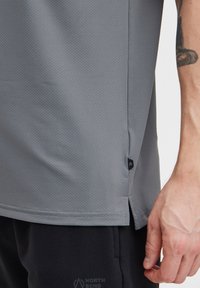 Šedé sportovní tričko s texturovaným povrchem, postranními rozparky a malou značkovou nášivkou na lemu. V kombinaci s černými šortkami.