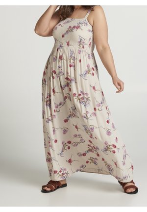 Nainen beige maxi-mekossa, jossa on punainen ja violetti kukkakuosi, ja ruskeat sandaalit, poseeraa yksinkertaista taustaa vasten.