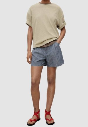 Beige katoenen t-shirt met opgerolde mouwen, gecombineerd met marineblauwe gestreepte korte broek. Model draagt rode sandalen met bandjes en een zwarte zool.