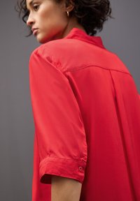 Camicia rossa a maniche corte con polsini abbottonati, caratterizzata da una texture liscia e lucida e un dettaglio sullo yoke posteriore, su uno sfondo grigio.
