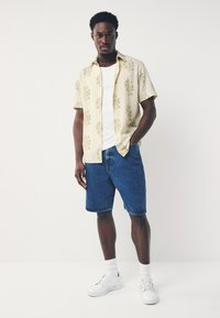 Camicia a maniche corte giallo chiaro con motivo floreale, abbinata a pantaloncini di denim blu e sneaker bianche. Lo sfondo è bianco uniforme.