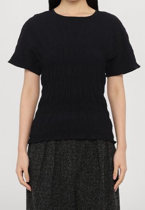 Haut noir à manches courtes, tissu texturé, design côtelé et encolure ronde. Contexte comprenant une jupe noire à motifs.