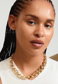 Collana in oro con maglie intrecciate e una finitura liscia e lucida. Indossata sopra una maglia leggera a coste, su uno sfondo neutro.