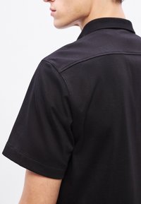 Μαύρο πόλο πουκάμισο με γιακά και κοντά μανίκια. Το ύφασμα έχει ομαλή υφή, με ραφές λεπτομέρειας στους ώμους.