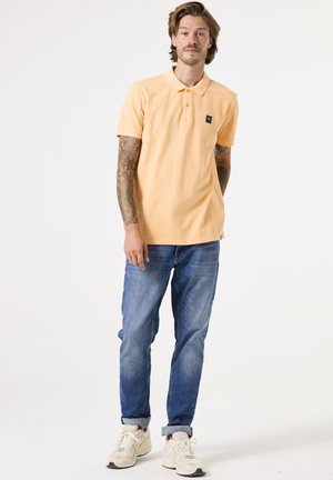 Man met tatoeages, gekleed in een perzikkleurig poloshirt, blauwe spijkerbroek en witte sneakers, staand tegen een witte achtergrond.