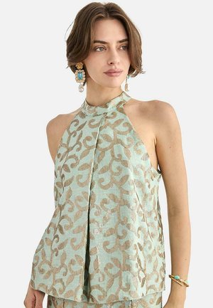 Joven con cabello corto castaño viste una blusa sin mangas de cuello alto color verde menta con patrones dorados en espiral y lleva pendientes grandes turquesa y de perlas.