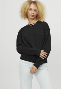 Felpa nera con collo a giro, spalle scese e polsini a costine. Indossata con jeans azzurri chiari. Tessuto liscio, design casual.