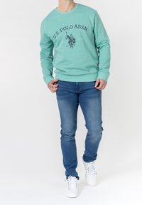 Man wearing teal U.S. Polo Assn. sweatshirt, blue jeans, and white sneakers, standing against white background.