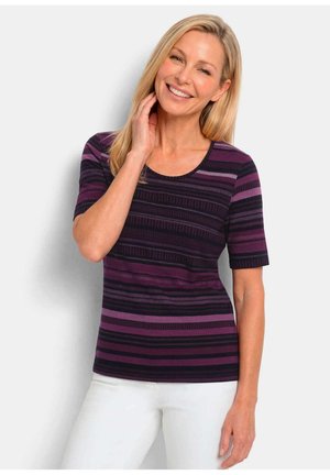 Femme souriante aux cheveux blonds portant un haut rayé violet et noir à manches courtes et un pantalon blanc, debout devant un fond uni.
