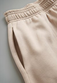 Béžové tepláky s žebrovaným pasem, bočními kapsami a měkkou, texturovanou tkaninou. Design zahrnuje čisté linie a vypasovaný střih.