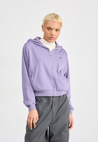 Felpa zip-up viola realizzata in misto cotone, con tasca frontale, polsini a coste e un piccolo logo nero sul petto. Indossata con pantaloni grigi.