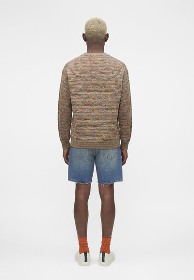 Άτομο με κοντά ξανθά μαλλιά που φοράει καφέ πολύχρωμο πλεκτό πουλόβερ, μπλε τζιν σορτς, πορτοκαλί κάλτσες και λευκά αθλητικά παπούτσια, στέκεται γυρισμένο με την πλάτη.