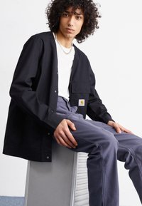 Black button-up shirt with a front pocket and Carhartt logo, paired with light grey pants, sitting on a metallic surface.