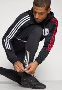 Chándal deportivo negro con rayas rojas y blancas. Pantalones ajustados y sudadera con capucha. Zapatillas de fútbol negras ajustables individualmente con superficie texturizada.