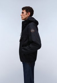 Ein Mann steht im Profil, trägt eine schwarze Kapuzenjacke mit einem Logopatch am Ärmel und blaue Jeans vor einem schlichten hellen Hintergrund.