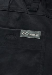 Κοντινό πλάνο μαύρης υφασμάτινης τσέπης με ορατή ραφές και γκρι ορθογώνια ετικέτα με το λογότυπο Columbia ραμμένη πάνω της.