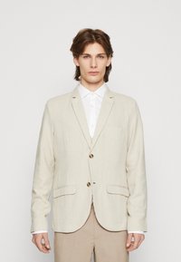 Ljust beige linneblazer med subtila vertikala ränder, två framfickor och en enkelknappslåsning. Bärs över en vit skjorta.