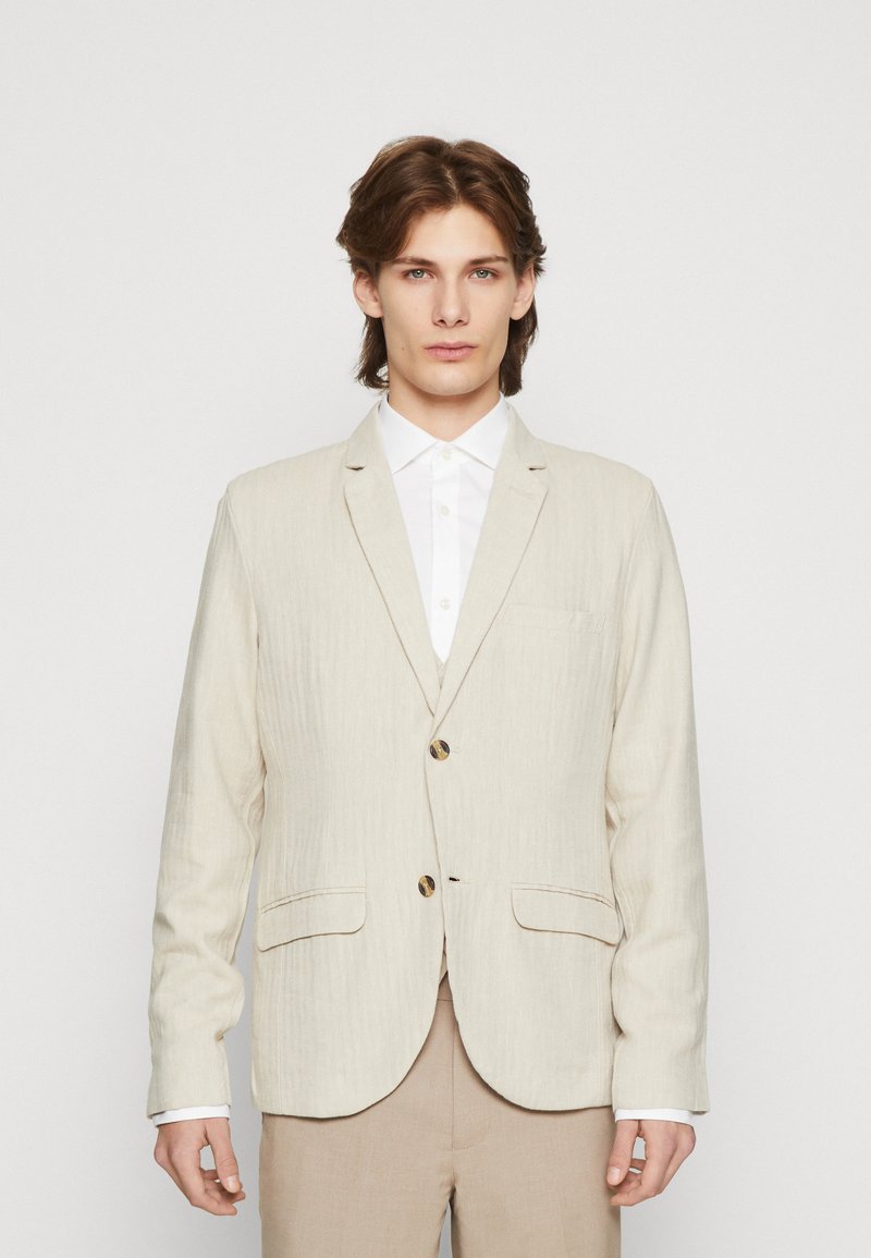 Ljust beige linneblazer med subtila vertikala ränder, två framfickor och en enkelknappslåsning. Bärs över en vit skjorta.