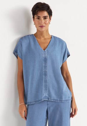 Femme aux cheveux courts et foncés, vêtue d'un haut en denim bleu clair à col en V et d'un pantalon assorti, debout devant un fond blanc, les mains détendues.