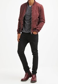 Mężczyzna w bordo bomberce, ciemnoszarej koszulce typu henley, czarnych wąskich spodniach i bordowych skórzanych butach typu Chelsea, stoi z rękami w kieszeniach.