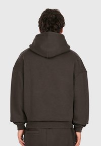 Uomo che indossa un ampio hoodie marrone scuro e pantaloni coordinati, visto da dietro su uno sfondo leggero e uniforme.