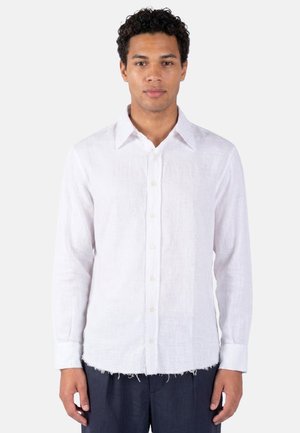 Uomo con capelli ricci che indossa una camicia bianca con bottoni e orlo sfrangiato e pantaloni scuri, in piedi davanti a uno sfondo chiaro e uniforme.