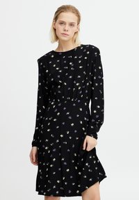 Robe noire à manches longues, arborant un motif floral subtil en violet et vert. Tissu texturé, taille cintrée, et forme évasée.
