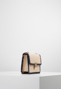 Borsa a tracolla in pelle beige con rifiniture ondulate blu navy, accessori in oro e una chiusura frontale. Forma rettangolare compatta. Texture liscia.