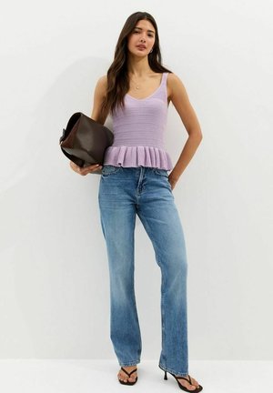 Donna che indossa un top senza maniche di maglia lilla con orlo a volant, jeans blu a vita alta, sandali neri con tacco, che tiene in mano una grande borsa marrone scuro.